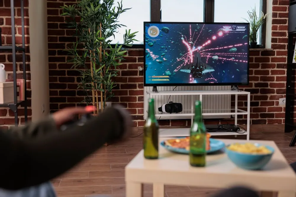 Salón con una televisión encendida en una partida de videojuego, un mando en primer plano y pizza y bebidas sobre la mesa